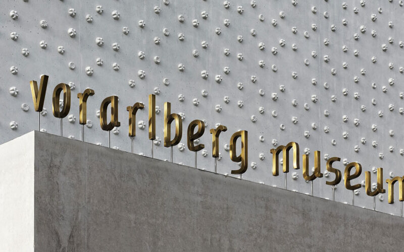 Goldene Buchstaben mit dem Schriftzug "vorarlberg museum" sind auf einer Betonwand mit einer strukturierten, metallischen Oberfläche angebracht, die im Hintergrund erhabene kreisförmige Muster aufweist.
