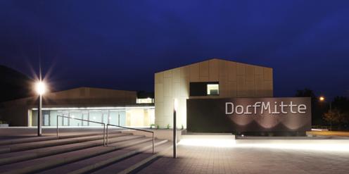 Modernes Gebäude mit der Aufschrift "DorfMitte", nachts beleuchtet, mit Treppen und einer Straßenlaterne im Vordergrund.