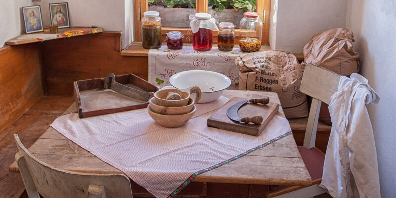 Eine rustikale Küche mit einem Holztisch, der mit Schüsseln, einem Schneidewerkzeug und einer Schüssel gedeckt ist; Gläser mit Eingemachtem und Getreidesäcke stehen in der Nähe eines sonnendurchfluteten Fensters.