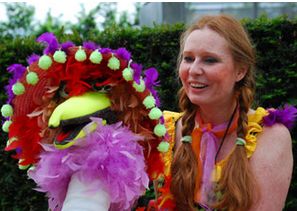 Eine Frau mit rotem Haar und farbenfroher Kleidung hält eine lebhafte, mit Federn und Bommeln geschmückte Puppe, die im Freien vor einer Grünanlage steht.