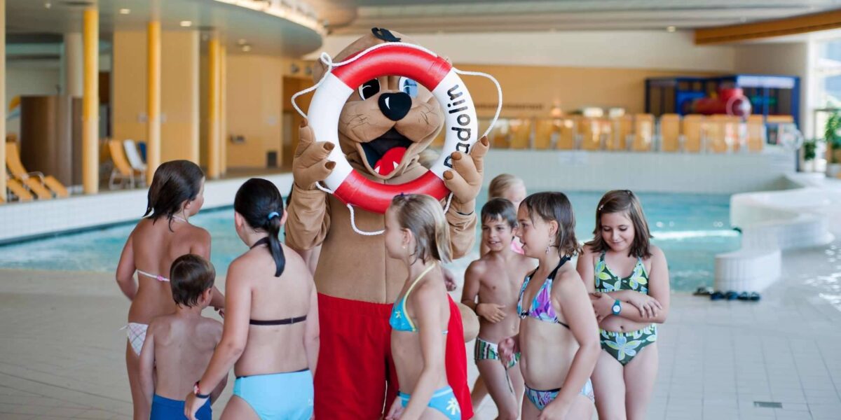 Eine Person in einem Tiermaskottchenkostüm mit einem Rettungsring steht neben einer Gruppe von Kindern in Badeanzügen in einem Hallenbad.