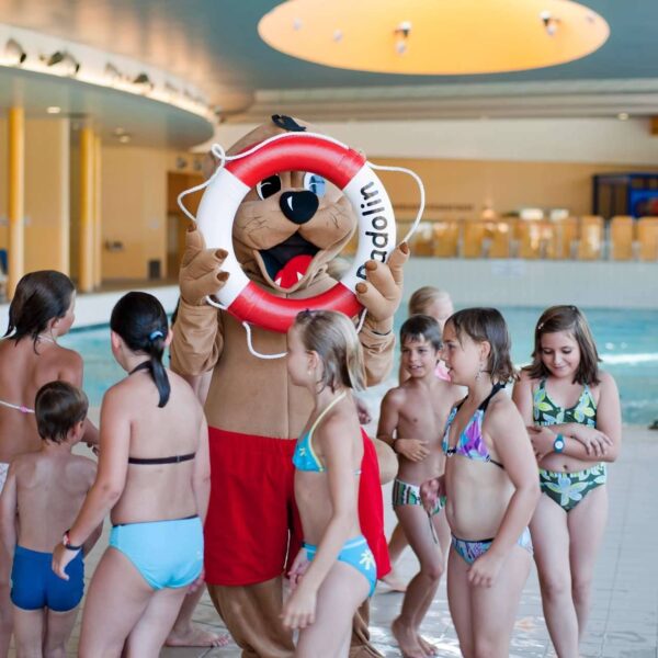 Eine Person in einem Tiermaskottchenkostüm mit einem Rettungsring steht neben einer Gruppe von Kindern in Badeanzügen in einem Hallenbad.