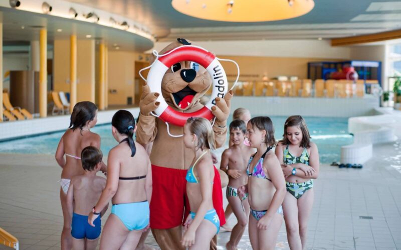 Eine Person in einem Tiermaskottchenkostüm mit einem Rettungsring steht neben einer Gruppe von Kindern in Badeanzügen in einem Hallenbad.