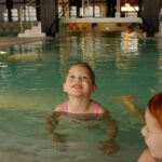 Zwei kleine Kinder befinden sich in einem Hallenbad. Ein Kind in einem rosafarbenen Badeanzug steht mit dem Gesicht nach vorne, und ein anderes mit roten Haaren ist teilweise im Vordergrund zu sehen.