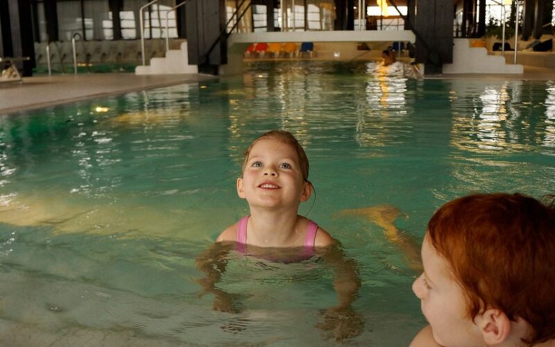 Zwei kleine Kinder befinden sich in einem Hallenbad. Ein Kind in einem rosafarbenen Badeanzug steht mit dem Gesicht nach vorne, und ein anderes mit roten Haaren ist teilweise im Vordergrund zu sehen.