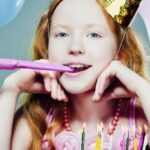 Junges Mädchen mit roten Haaren, das einen Partyhut trägt und in ein Partyhorn bläst, sitzt vor einer Geburtstagstorte mit brennenden Kerzen und ist von Luftballons umgeben.