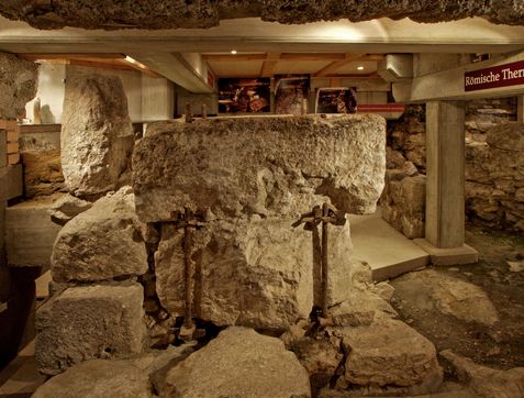 Große, verwitterte Steinstrukturen und Säulen werden in einer überdachten archäologischen Stätte mit Oberlicht und einem Schild mit der Aufschrift "Römische Thermen" ausgestellt.