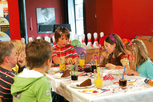Kinder sitzen um einen geschmückten Tisch herum, essen Kuchen und trinken Saft auf einer Geburtstagsfeier, mit Luftballons und Bowlingkegeln im Hintergrund.
