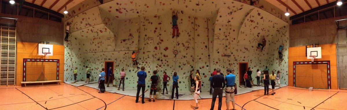 Eine Gruppe von Personen in einer Sporthalle, in der mehrere Personen eine große Kletterwand erklimmen, während andere zusehen oder warten, bis sie an der Reihe sind.