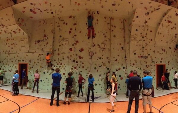 Eine Gruppe von Personen in einer Sporthalle, in der mehrere Personen eine große Kletterwand erklimmen, während andere zusehen oder warten, bis sie an der Reihe sind.