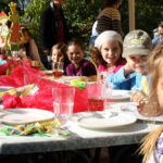 Eine Gruppe von Kindern sitzt um einen geschmückten Tisch im Freien und lächelt in die Kamera, mit Tellern, Gläsern und buntem Partyschmuck im Sonnenlicht.