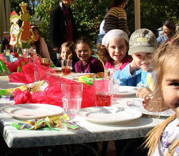 Eine Gruppe von Kindern sitzt um einen geschmückten Tisch im Freien und lächelt in die Kamera, mit Tellern, Gläsern und buntem Partyschmuck im Sonnenlicht.