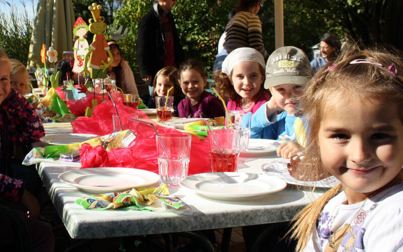 Eine Gruppe von Kindern sitzt um einen geschmückten Tisch im Freien und lächelt in die Kamera, mit Tellern, Gläsern und buntem Partyschmuck im Sonnenlicht.