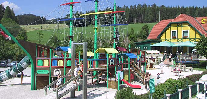 Spielplatz im Freien mit einem großen Piratenschiff mit Rutschen, Kletternetzen und spielenden Kindern vor einer Kulisse aus Bäumen und einem Gebäude mit rotem Dach.