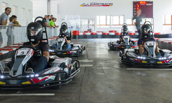Drei Personen mit Helmen fahren Go-Karts auf einer Indoor-Rennstrecke, während Zuschauer hinter einer Absperrung zuschauen. Im Hintergrund ist eine digitale Zeitmessanlage zu sehen.