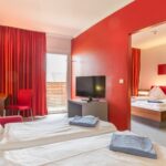 Zwei Hotelzimmerbetten mit weißer Bettwäsche und gefalteten Handtüchern, ein Fernseher, ein Schreibtisch mit einer Lampe, rote Wände, rote Vorhänge und ein Verbindungszimmer mit einem weiteren Doppelbett.