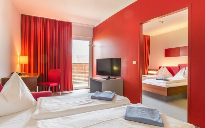 Zwei Hotelzimmerbetten mit weißer Bettwäsche und gefalteten Handtüchern, ein Fernseher, ein Schreibtisch mit einer Lampe, rote Wände, rote Vorhänge und ein Verbindungszimmer mit einem weiteren Doppelbett.