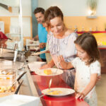 Eine Frau und ein junges Mädchen servieren das Essen auf Tellern in einer Cafeteria, während ein Mann hinter ihnen in der Schlange wartet.