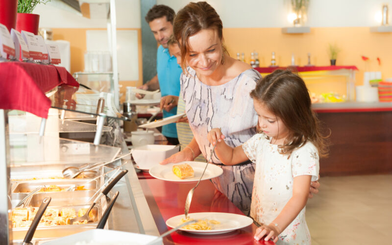 Eine Frau und ein junges Mädchen servieren das Essen auf Tellern in einer Cafeteria, während ein Mann hinter ihnen in der Schlange wartet.