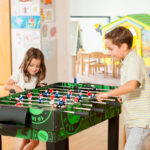 Zwei Kinder spielen Tischfußball an einem grünen Tisch in einem hell erleuchteten Raum mit Kinderzeichnungen an den Wänden und einem gelben Spielhaus im Hintergrund.