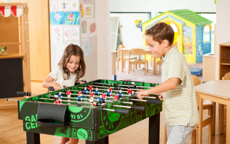 Zwei Kinder spielen Tischfußball an einem grünen Tisch in einem hell erleuchteten Raum mit Kinderzeichnungen an den Wänden und einem gelben Spielhaus im Hintergrund.