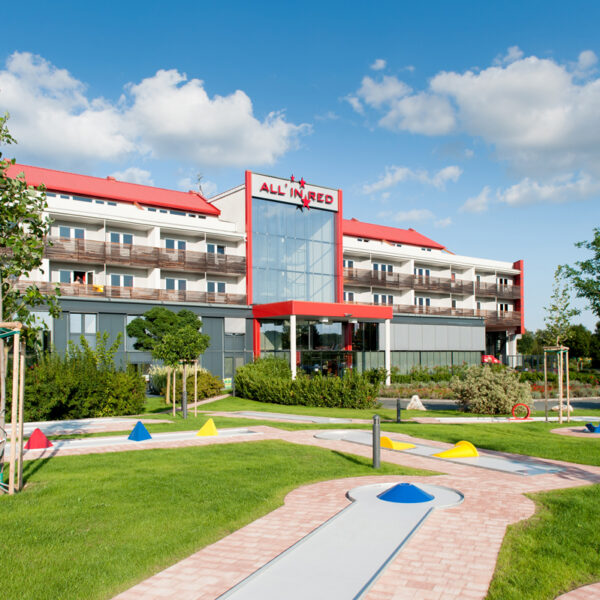Ein modernes Hotelgebäude mit roten Akzenten und Balkonen, mit einem begrünten Minigolfplatz im Vordergrund unter einem teilweise bewölkten Himmel.