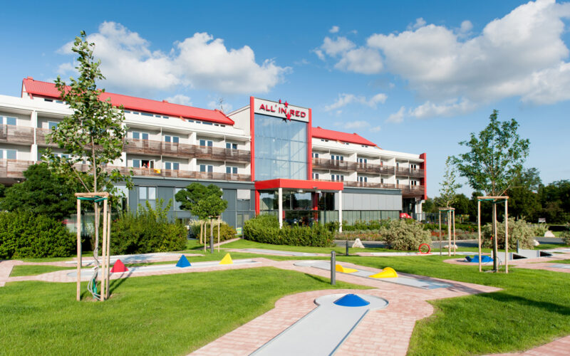 Ein modernes Hotelgebäude mit roten Akzenten und Balkonen, mit einem begrünten Minigolfplatz im Vordergrund unter einem teilweise bewölkten Himmel.