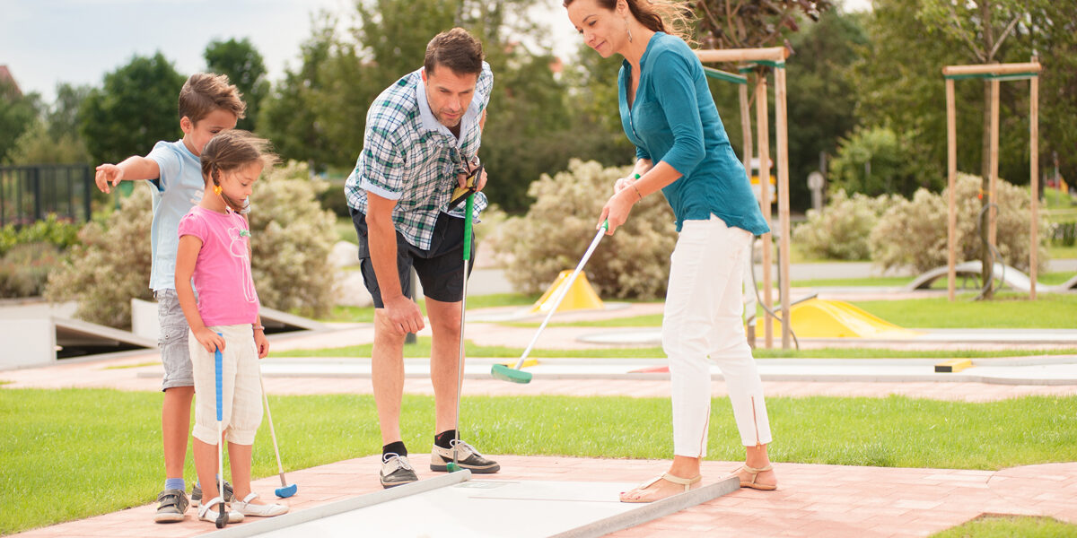 Eine vierköpfige Familie spielt im Freien Minigolf; die Eltern und zwei Kinder stehen mit Golfschlägern in der Hand auf dem Platz und konzentrieren sich auf das Spiel.