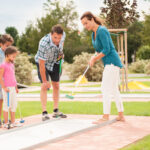 Eine vierköpfige Familie spielt im Freien Minigolf; die Eltern und zwei Kinder stehen mit Golfschlägern in der Hand auf dem Platz und konzentrieren sich auf das Spiel.