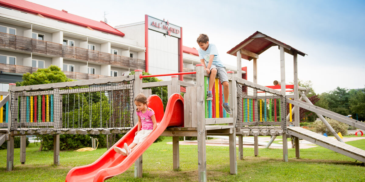 Zwei Kinder spielen auf einem hölzernen Spielplatz; eines rutscht eine rote Rutsche hinunter, während das andere obenauf klettert. Im Hintergrund ist ein Gebäude mit einem roten Dach zu sehen.