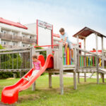 Zwei Kinder spielen auf einem hölzernen Spielplatz; eines rutscht eine rote Rutsche hinunter, während das andere obenauf klettert. Im Hintergrund ist ein Gebäude mit einem roten Dach zu sehen.