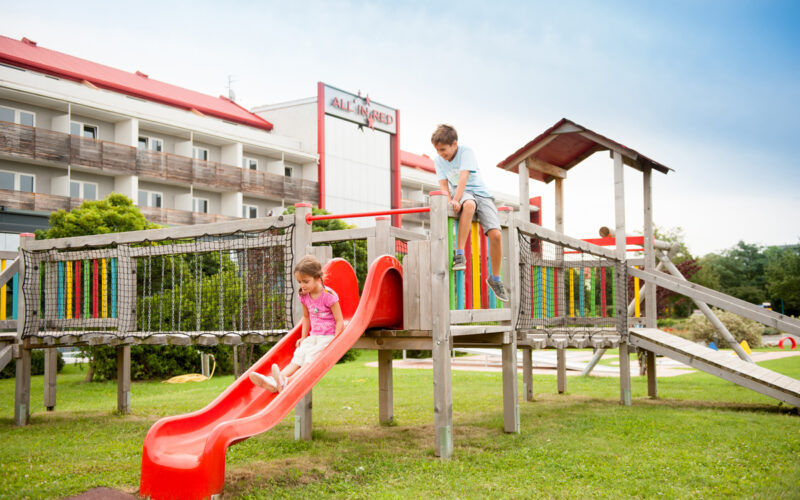 Zwei Kinder spielen auf einem hölzernen Spielplatz; eines rutscht eine rote Rutsche hinunter, während das andere obenauf klettert. Im Hintergrund ist ein Gebäude mit einem roten Dach zu sehen.