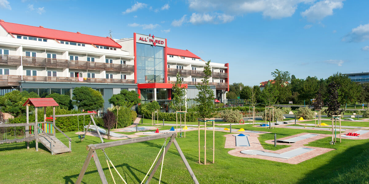 Ein Hotel mit rotem Dach und Balkonen überblickt an einem sonnigen Tag einen angelegten Spielplatz und eine Minigolfanlage.