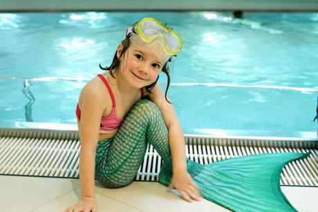 Ein kleines Kind in einem rosa Badeanzug, einem grünen Meerjungfrauenschwanz und einer gelben Schwimmbrille sitzt am Rand eines Hallenbades und lächelt in die Kamera.