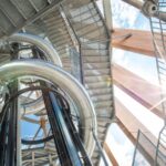 Blick von unten auf eine metallene Wendelrutsche und Treppen in einem Aussichtsturm aus Stahl und Holz, wobei das Sonnenlicht durch die Struktur scheint.