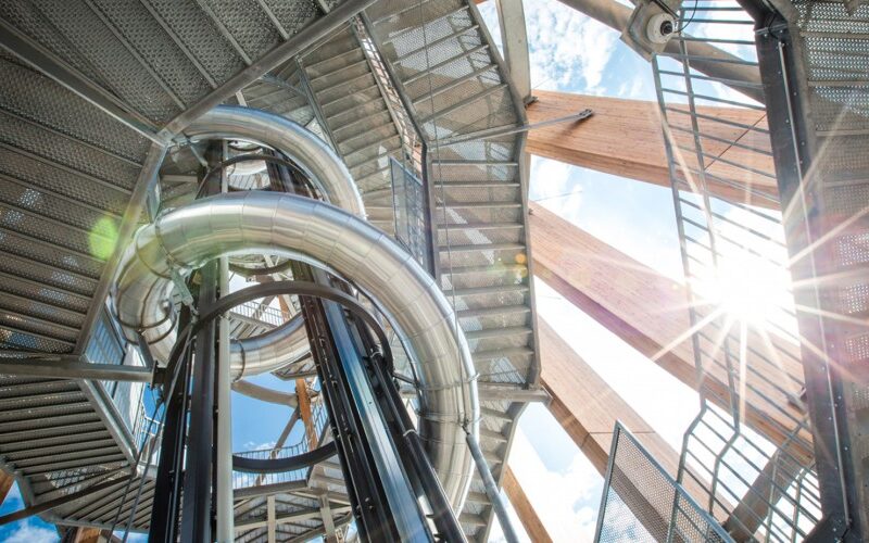 Blick von unten auf eine metallene Wendelrutsche und Treppen in einem Aussichtsturm aus Stahl und Holz, wobei das Sonnenlicht durch die Struktur scheint.