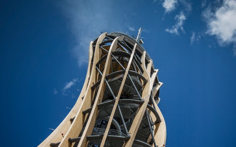 Ein hoher, spiralförmiger hölzerner Aussichtsturm mit Metallstützen steht vor einem klaren blauen Himmel mit ein paar Wolken.