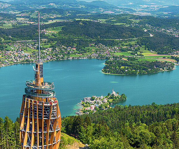Von einem hohen hölzernen Aussichtsturm aus überblickt man einen See, der von bewaldeten Hügeln, kleinen Städten und grünen Feldern umgeben ist.