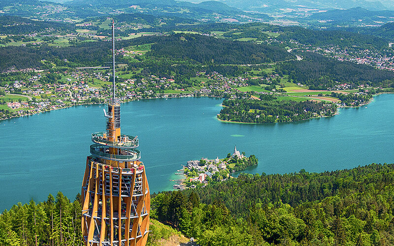 Von einem hohen hölzernen Aussichtsturm aus überblickt man einen See, der von bewaldeten Hügeln, kleinen Städten und grünen Feldern umgeben ist.
