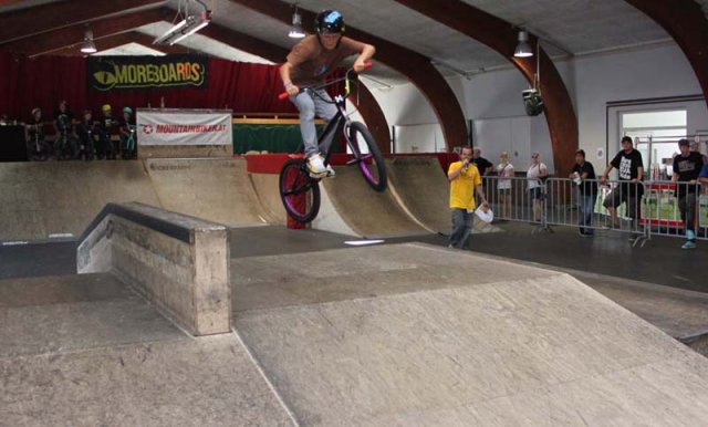 Ein BMX-Fahrer führt in einem Indoor-Skatepark einen Trick in der Luft aus, während die Zuschauer hinter einem Geländer zuschauen.