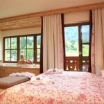 Ein gemütliches Schlafzimmer mit geblümter Tagesdecke, großen Fenstern und einer Person, die in einer Wanne badet, von der aus man einen schönen Blick auf die Berge hat.