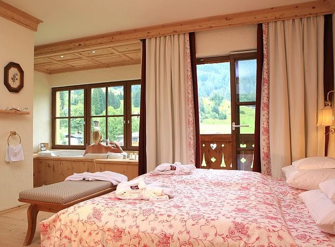 Ein gemütliches Schlafzimmer mit geblümter Tagesdecke, großen Fenstern und einer Person, die in einer Wanne badet, von der aus man einen schönen Blick auf die Berge hat.