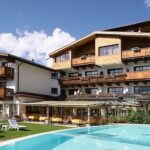 Ein Hotel mit Holzbalkonen und einem schrägen Dach steht hinter einem klaren Außenpool und Liegestühlen an einem sonnigen Tag.