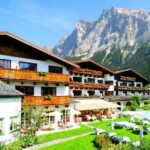 Ein großes Alpenhotel mit Holzbalkonen und Blick auf einen Garten und einen Außenpool vor der Kulisse hoher Berge und des blauen Himmels.