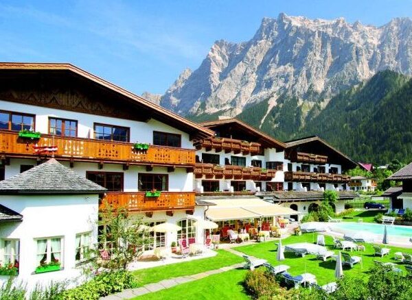 Ein großes Alpenhotel mit Holzbalkonen und Blick auf einen Garten und einen Außenpool vor der Kulisse hoher Berge und des blauen Himmels.