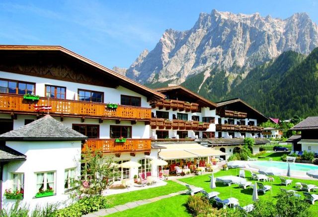 Ein großes Alpenhotel mit Holzbalkonen und Blick auf einen Garten und einen Außenpool vor der Kulisse hoher Berge und des blauen Himmels.