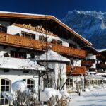 Eine schneebedeckte Berghütte mit Holzbalkonen liegt am Fuße eines Berges unter einem strahlend blauen Himmel.