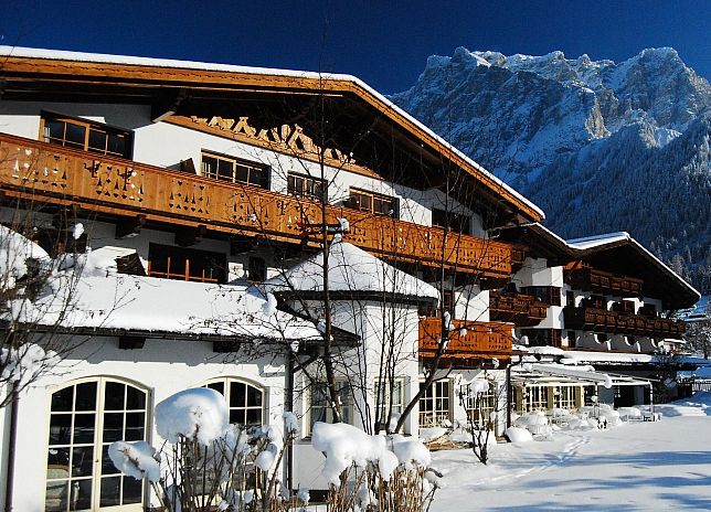Eine schneebedeckte Berghütte mit Holzbalkonen liegt am Fuße eines Berges unter einem strahlend blauen Himmel.