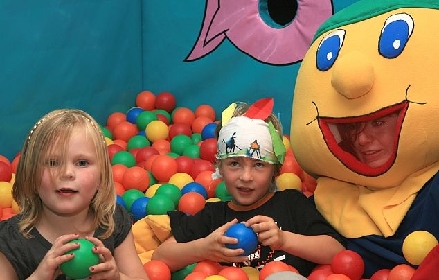 Drei Kinder sitzen in einem bunten Bällebad, eines trägt ein Stirnband mit Zeichnungen, ein anderes ein Figurenkostüm mit einem großen lächelnden Gesicht.