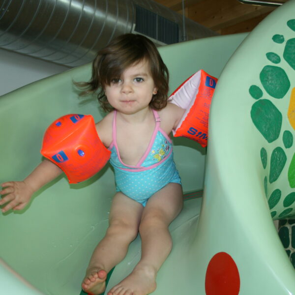 Ein kleines Kind, das orangefarbene Schwimmflügel und einen Badeanzug trägt, rutscht eine grüne Indoor-Wasserrutsche mit einem Schildkrötenmosaik hinunter.